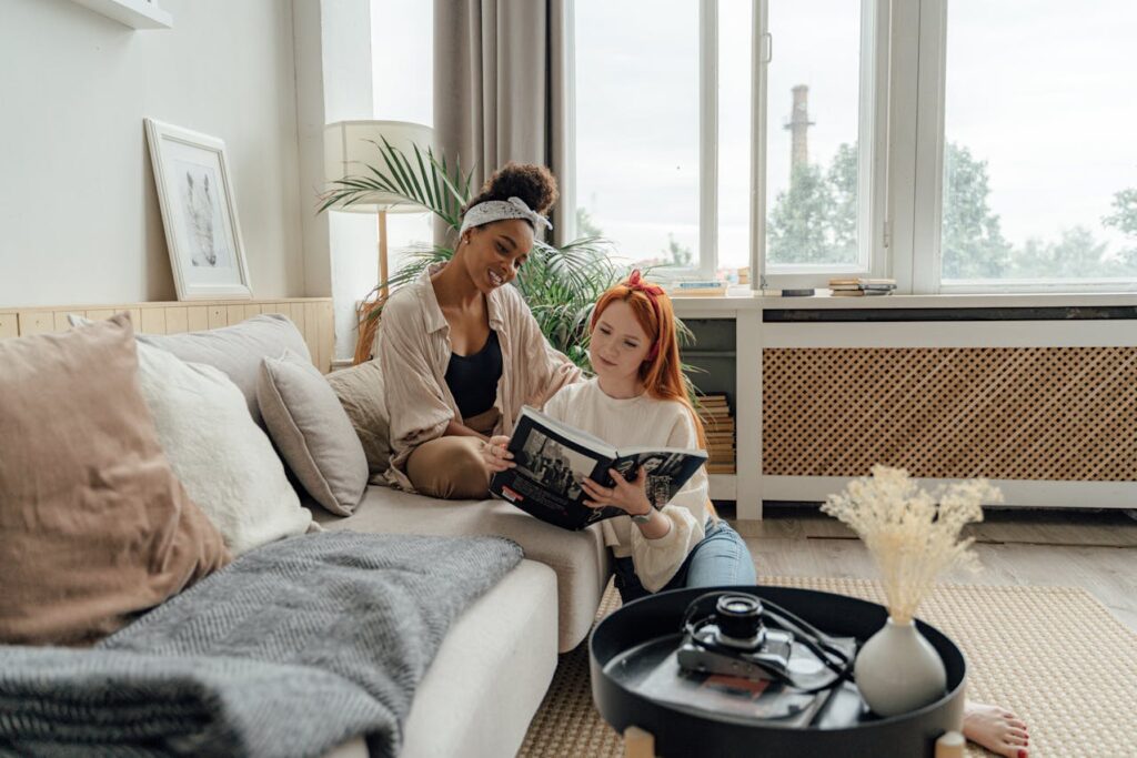 Two people looking at a book in a bright living room with large windows in the rear