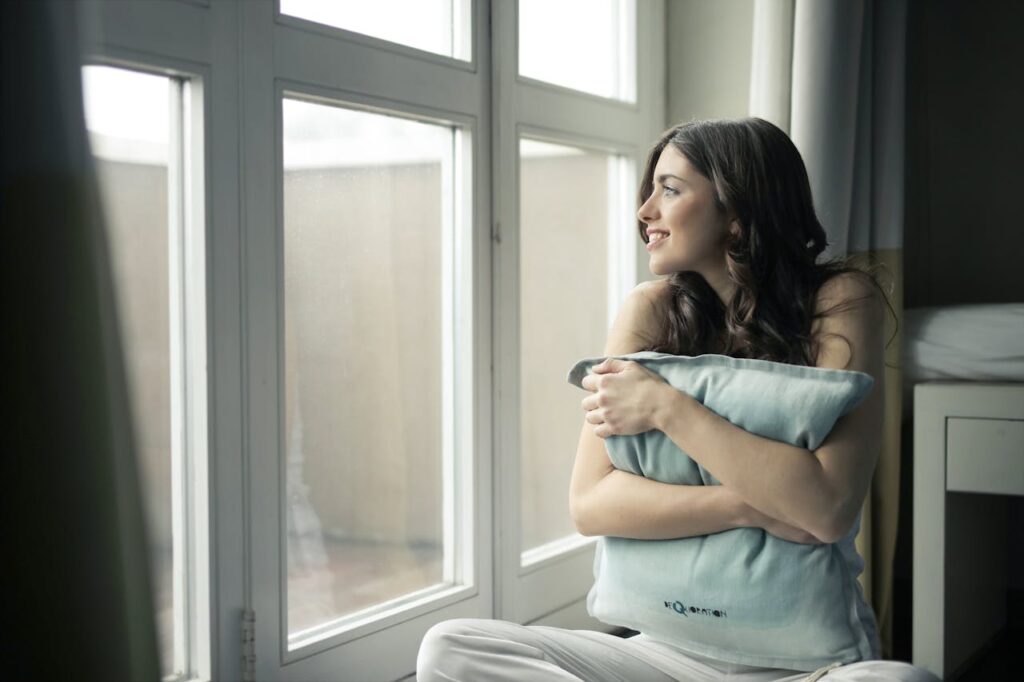 Smiling person sitting in front of windows, looking outward and hugging pillow